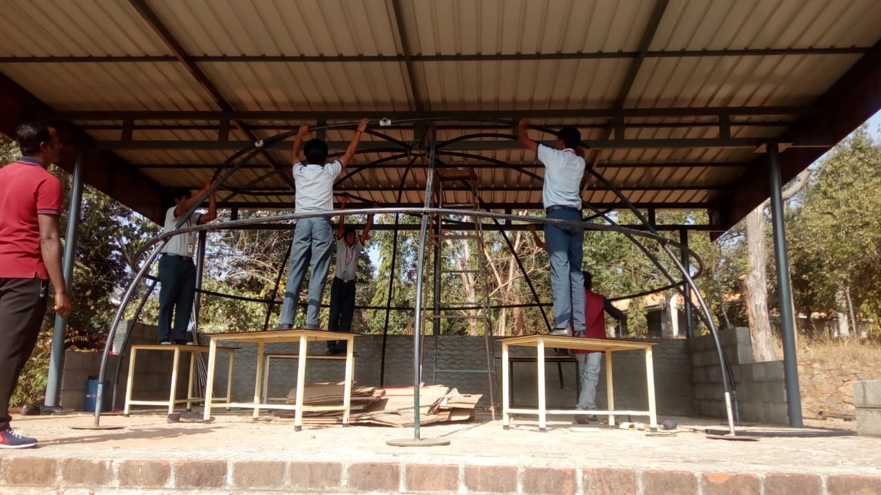 Students and a teacher assembling the bare geodesic frame of the Version 2 planetarium dome, standing on desks to reach the high ribs