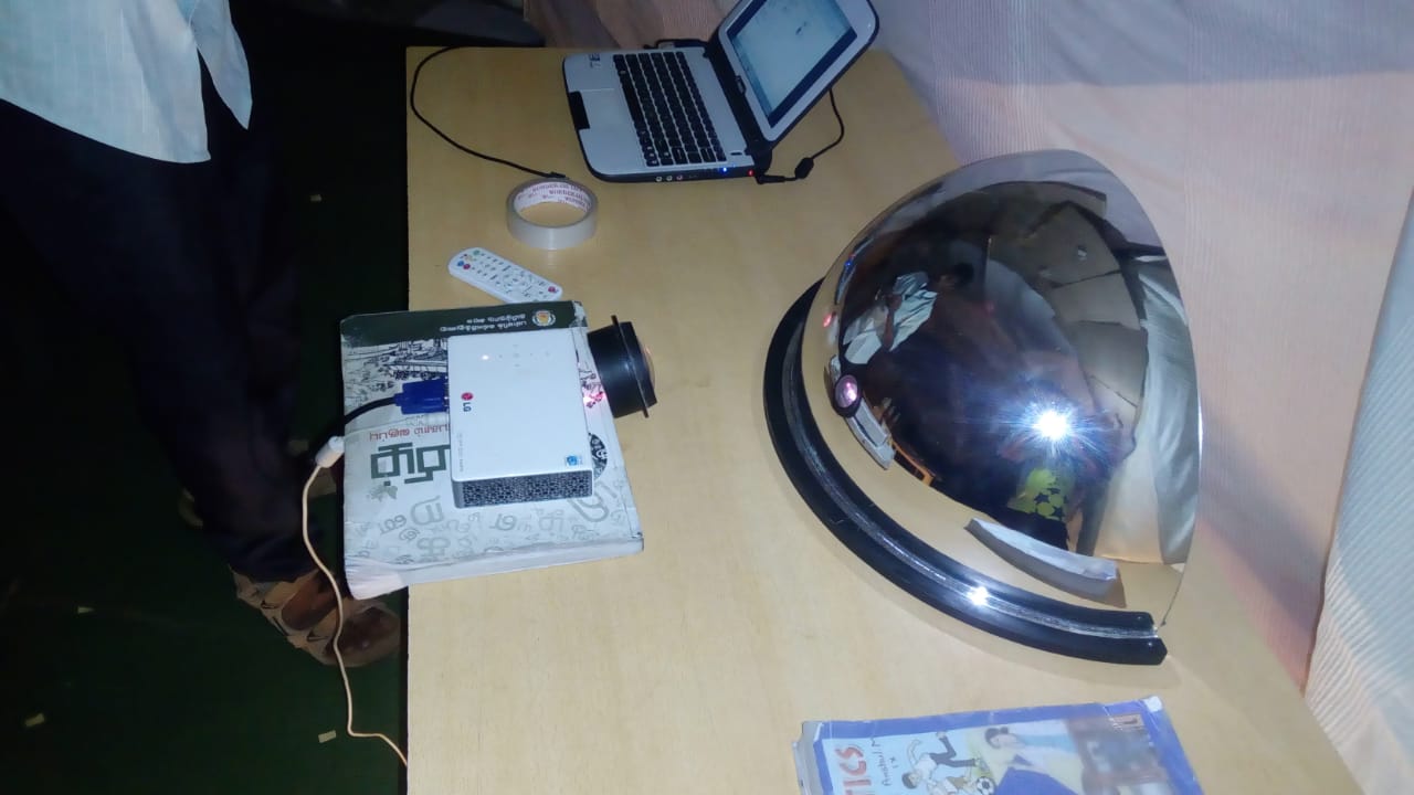 A laptop, projector, and convex spherical mirror set up on a table inside the planetarium, with a physics textbook alongside