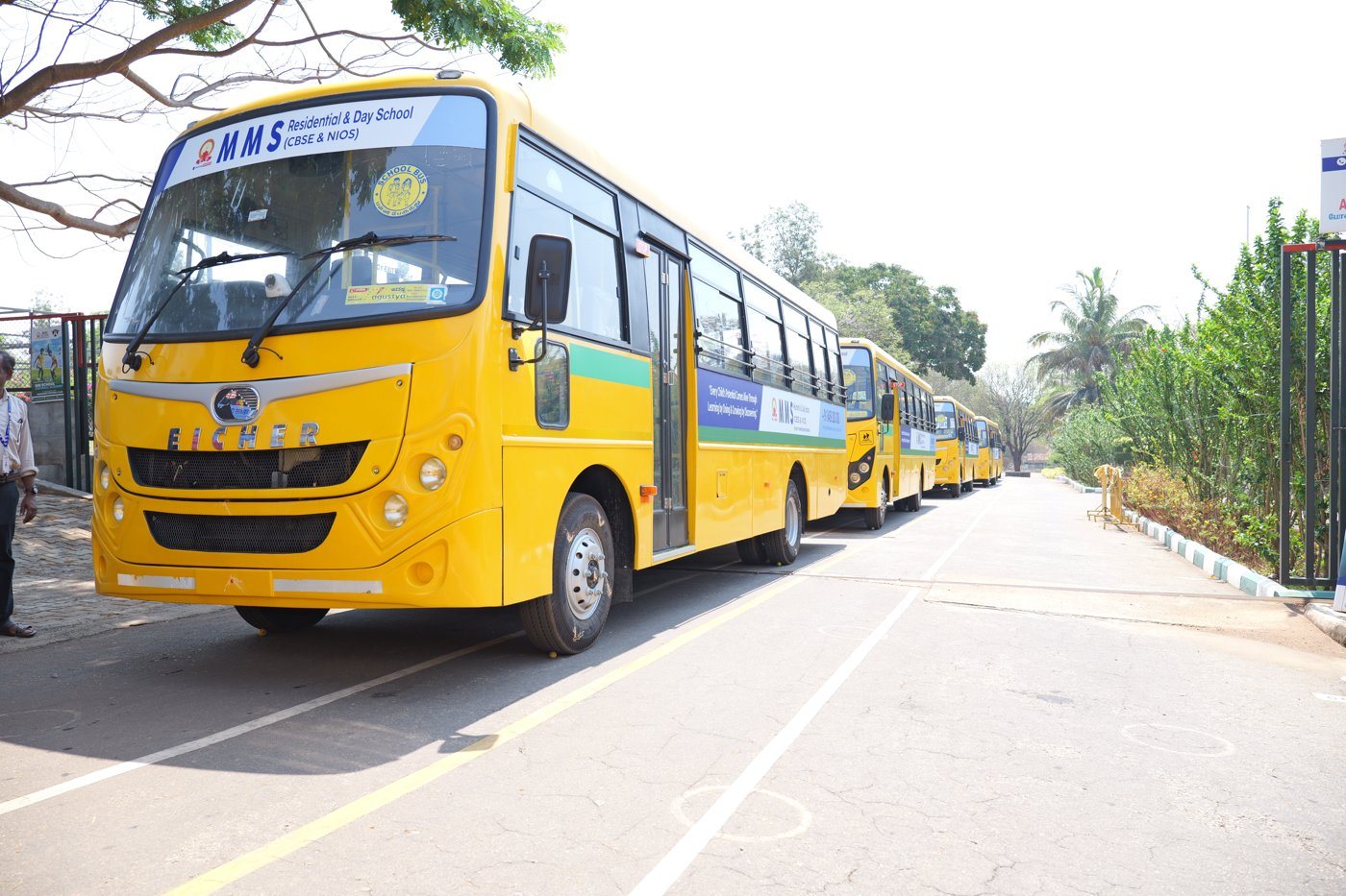A new yellow MMS school bus at the campus road, with the rest of the fleet lined up behind it